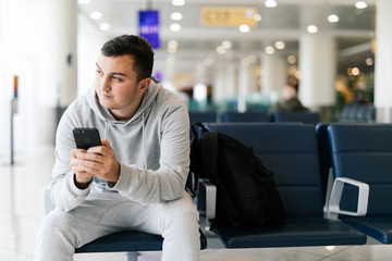 Texting he is fine. Portrait of handsome smiling man in casual wear holding his luggage and messaging through his mobile phone while sitting in the hall of the airport