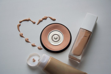 Beige dashed heart with dry and liquid skin tone correctors: compact powder, tonal cream in glass bottle and plastic tube on white background