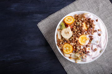 Vegetarian salad of bananas, apples, pears, kumquats, kiwi with granola and yogurt on black wooden background.