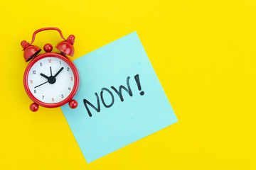 Sticky post with handwriting the word Now with red alarm clock on solid yellow background with copy...