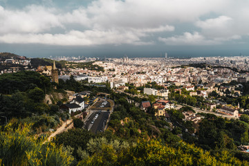 Beautiful evening scenery of Barcelona, Spain: the cityscape with many famous buildings, different districts, and houses, the sea in the distance; with hills, greenery, road and park in the foreground