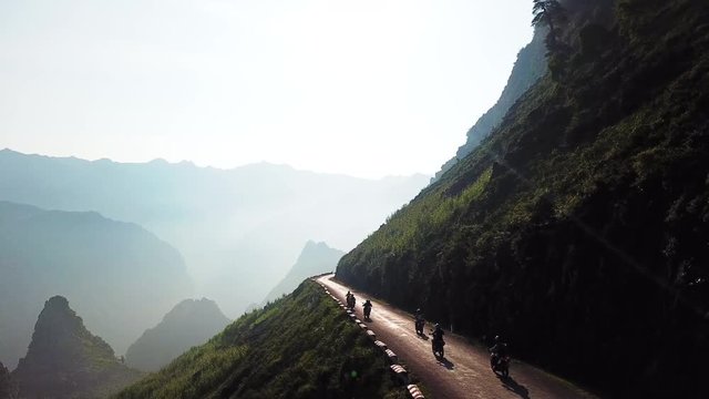 Aerial view alongside a mountainside, travelling with line of motorcyclists on the Ma Pi Leng Pass, mountain peaks in the foggy distance.
