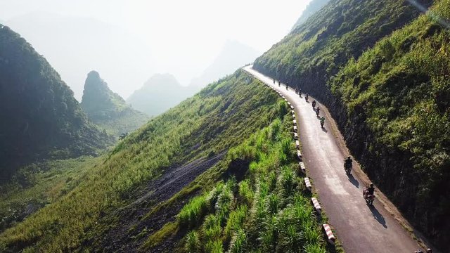 Drone view alongside a mountainside, travelling with line of motorcyclists on the Ma Pi Leng Pass, mountain peaks in the distance.