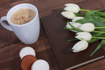 Bunch of white tulips, book in dark cover, cup of coffee and dessert on brown  wooden table. Holiday concept. Spring flowers, cappuccino, coffee break, good morning