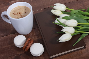 Bunch of white tulips, book in dark cover, cup of coffee and dessert on brown  wooden table. Holiday concept. Spring flowers, cappuccino, coffee break, good morning