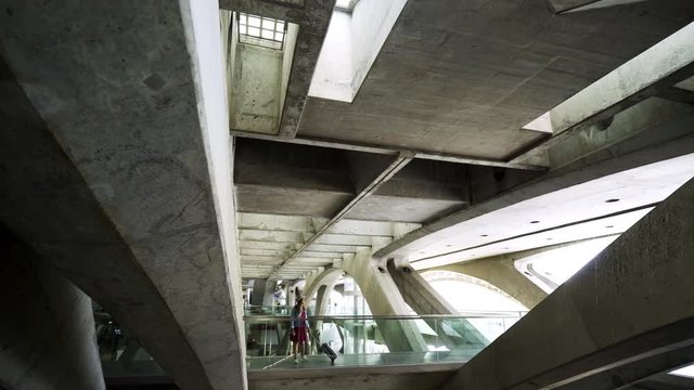 Young female passenger in stylish outfit pulling suitcase while walking between concrete and glass construction inside modern transportation terminal