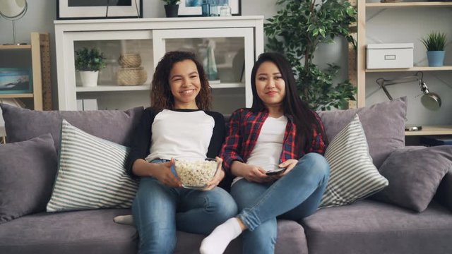 Happy female friends are watching funny movie together laughing, talking and eating popcorn sitting on couch at home. Friendship and television concept.