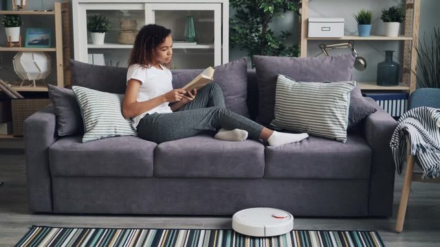 Relaxed African American girl is reading book resting on sofa while robotic vacuum cleaner is hoovering floor in house instead of person. Gadgets and household concept.