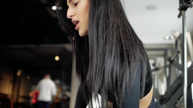 Attractive young caucasian girl doing biceps exercises while sitting on the bench and raising dumbbell in the gym