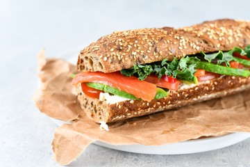 sandwich from a cereal baguette with avocado, salmon, cream cheese, tomatoes and lettuce leaves on a white plate.  light background, selective focus and copy space.