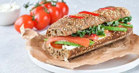 sandwich from a cereal baguette with avocado, salmon, cream cheese, tomatoes and lettuce leaves on a white plate.  light background, selective focus and copy space.