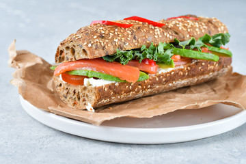 sandwich from a cereal baguette with avocado, salmon, cream cheese, tomatoes and lettuce leaves on a white plate.  light background, selective focus and copy space.