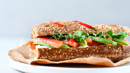 sandwich from a cereal baguette with avocado, salmon, cream cheese, tomatoes and lettuce leaves on a white plate.  light background, selective focus and copy space.