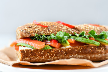sandwich from a cereal baguette with avocado, salmon, cream cheese, tomatoes and lettuce leaves on a white plate.  light background, selective focus and copy space.