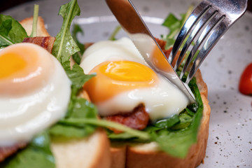 A woman eating breakfast sandwich with eggs, bacon and sour cream by knife and spoon in a plate on wooden table