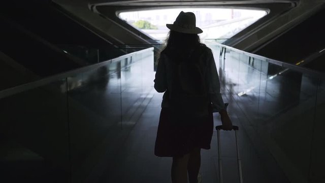 Back view of unrecognizable woman pulling suitcase and looking away while walking in dark passage inside modern railway station