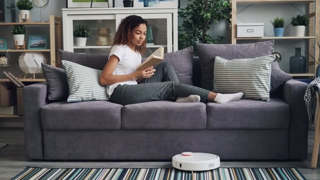 Good-looking African American student is turning on robotic hoover and reading book enjoying hobby and relaxing on couch while gadget is cleaning floor.
