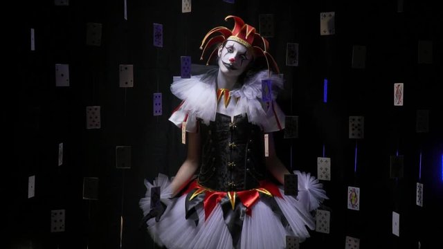 A girl jester is standing with her head tilted, playing cards are floating around, slow motion