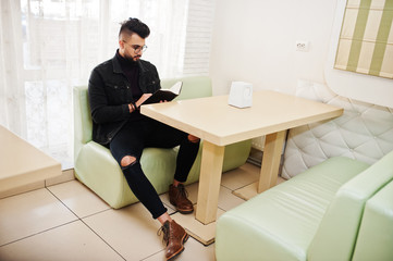 Arab man wear on black jeans jacket and eyeglasses sitting in cafe, read book. Stylish and fashionable arabian model guy.