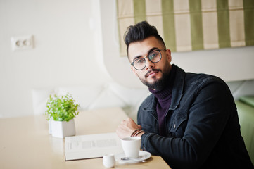 Arab man wear on black jeans jacket and eyeglasses sitting in cafe, read book and drink coffee. Stylish and fashionable arabian model guy.