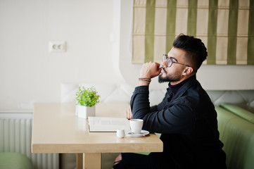 Arab man wear on black jeans jacket and eyeglasses sitting in cafe, read book and drink coffee. Stylish and fashionable arabian model guy.