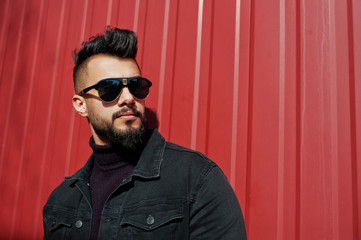 Fashion Arab man wear on black jeans jacket and sunglasses posed against red steel wall background....