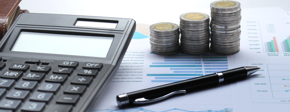 calculator, pile of coins, financial graph and pen on office desk, stock chart in background