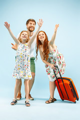 Fototapeta na wymiar Happy surprised parents with daughter and suitcase at studio isolated on blue background. Travel, vacation, parenthood, togetherness, tourism concept.