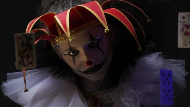 Close up of a creepy clown with makeup and old hat with bells, cards are floating in the air
