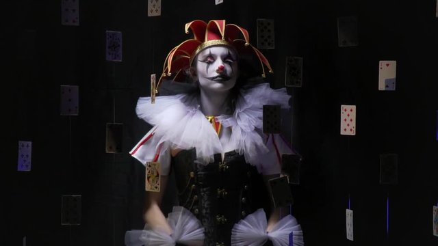 Female joker with a red hat and black and white dress is dancing among the floating in the air cards