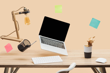 laptop with blank screen, lamp, empty sticky notes and stationery levitating in air above wooden desk with thermomug with coffee splash isolated on beige