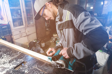 Young carpenter with a beard working with an electric plane with suction of sawdust. Leveling and sanding wooden bars