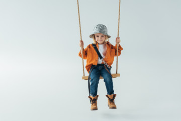 cute kid in silver hat, jeans and orange shirt sitting on swing on grey background - obrazy, fototapety, plakaty