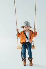 smiling kid in jeans and orange shirt sitting on swing on grey background