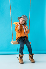 cute kid sitting on swing, holding paper plane and looking away on blue background