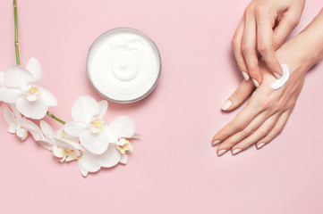 Young woman moisturizes her hand with cosmetic cream lotion opened container with cream body milk White Phalaenopsis orchid flowers on pink background Flat lay top view minimalism style Beauty concept