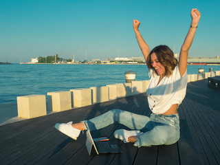 Excited young woman celebrating a triumph watching the laptop sitting on the bench