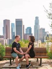 A young couple, a blond man and a brunette girl in a dress are sitting and chatting on a bench overlooking downtown Los Angeles