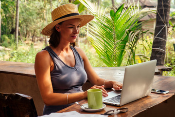 Technology and travel. Working outdoors. Freelance concept. Pretty young woman in hat using laptop in cafe on tropical beach.Hooray victory success and successful deal concept promotion at work
