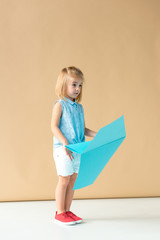 surprised  kid in shirt and shorts holding blue paper plane on beige background