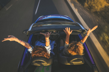 Top view a pair of euphoric women in a convertible car twisting and waving. Two curly girls on vacation having fun driving the auto, laughing happily singing, enjoying freedom in a beautiful sunlight - obrazy, fototapety, plakaty