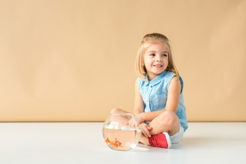 cute kid sitting on floor with fishbowl and looking away on beige background