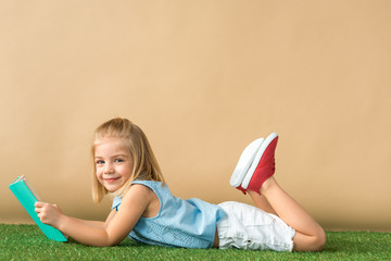 smiling and cute child lying on grass rug and holding book on beige background
