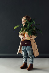  cute child in trench coat and jeans holding plant in flowerpot on black background