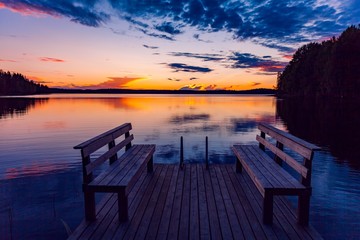 Fototapeta na wymiar Two wooden bench or chairs on a wood dock facing a lake at sunset in Finland