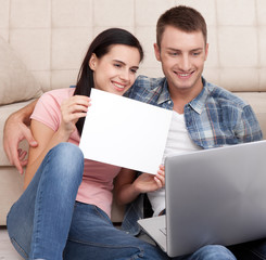Beautiful young couple using laptop communicates in video chat. A woman is smiling and showing blank piece of paper.