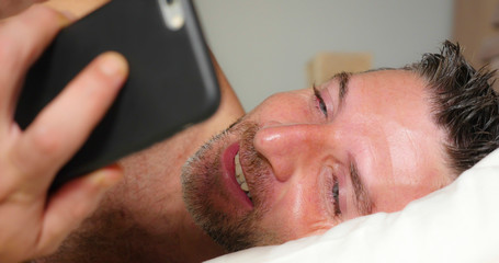 lifestyle portrait of young attractive and happy man lying on bed at home bedroom using mobile phone enjoying internet social media app or online gambling and dating