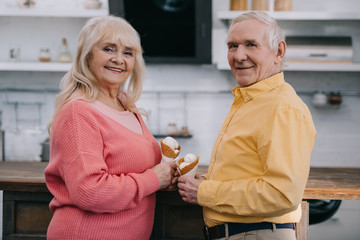 smiling senior couple looking at camera and holding ice cream cones at home