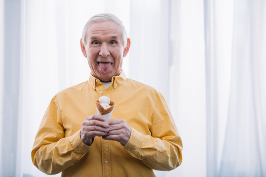 Senior Man Sticking Tongue Out, Looking At Camera And Holding Ice Cream Cone At Home With Copy Space