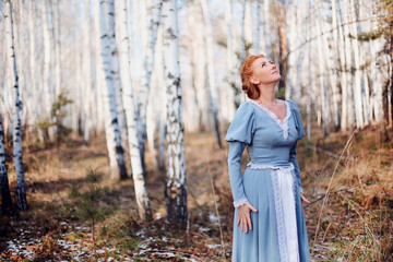 Beautiful young girl of European appearance with red hair and blue eyes on the background of spring birch forest. Happy woman  looking at the sky in nature. Illustration to a book or a fairy tale.